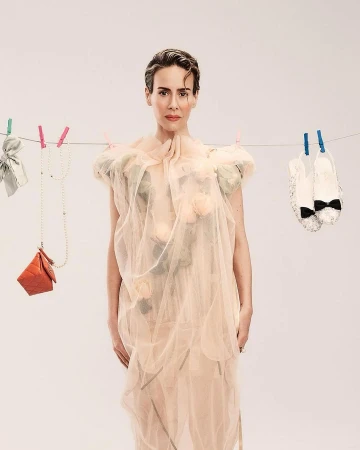 Woman in sheer gown poses in a photo studio, surrounded by fashion accessories hanging from clothespins and string against a light backdrop.