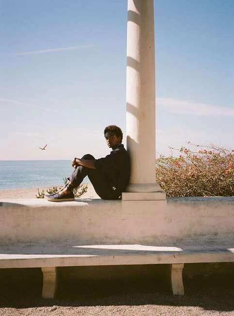 Young Black man sitting on a concrete ledge leaning against a pillar by the ocean, photographed by Victoria Will.