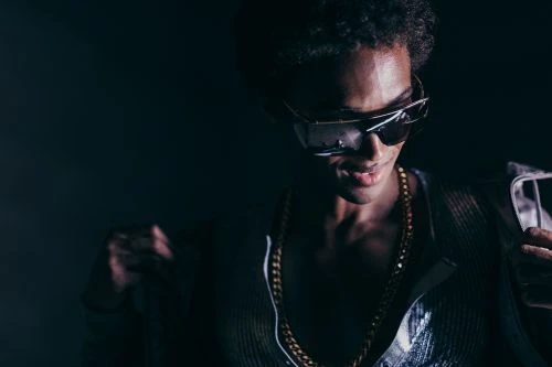 Portrait of a Black man wearing oversized sunglasses and a gold chain necklace under low-key dramatic lighting.
