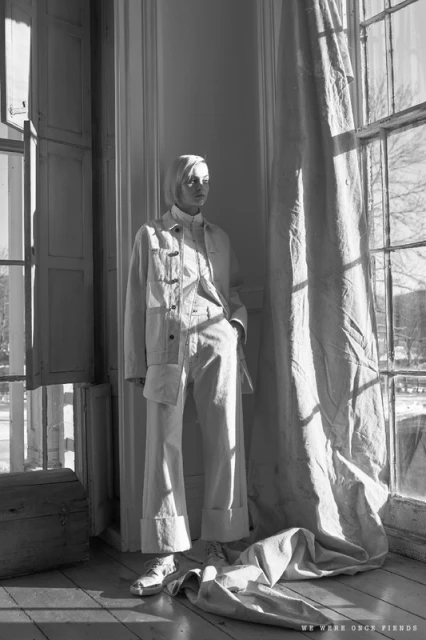 Monochrome fashion shot of a woman in a white jacket and pants standing near a window with light filtering through curtains.