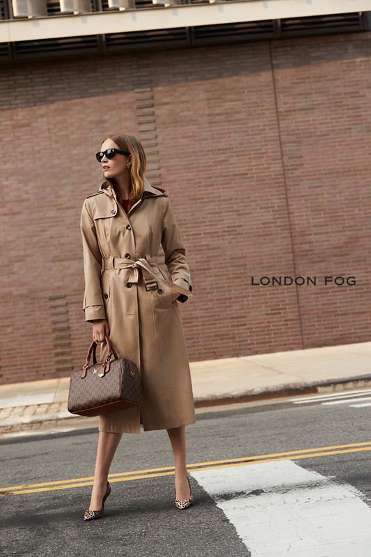A blonde woman poses in a trench coat with sunglasses, holding a purse in an urban setting with a brick building as backdrop.