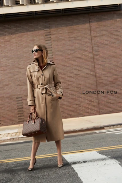 A blonde woman poses in a trench coat with sunglasses, holding a purse in an urban setting with a brick building as backdrop.