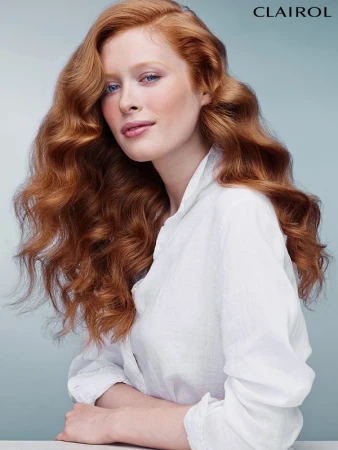 Red-haired woman with fair skin and light eyes in a white top posing against a gradient blue background, promoting hair color.