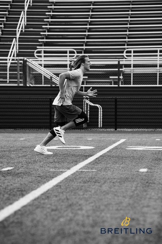 Black and white shot of a male athlete running on a football field with empty stadium seating in the background and a Breitling logo.