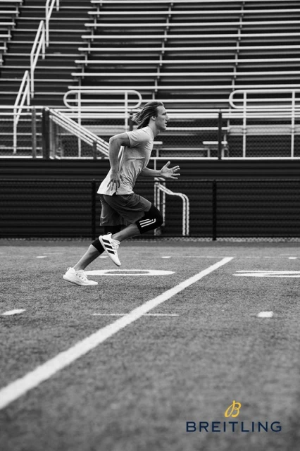 Black and white shot of a male athlete running on a football field with empty stadium seating in the background and a Breitling logo.