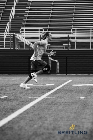 Black and white shot of a male athlete running on a football field with empty stadium seating in the background and a Breitling logo.