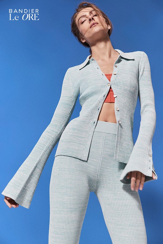 Woman in a light green knit outfit with flared sleeves poses against a bright blue backdrop, showcasing a modern and stylish fashion look.