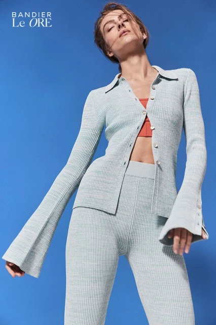 Woman in a light green knit outfit with flared sleeves poses against a bright blue backdrop, showcasing a modern and stylish fashion look.