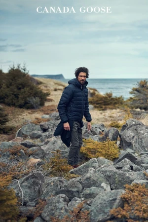 Man in a black puffer jacket poses on a rocky coastline with shrubs beneath a cloudy sky. Canada Goose logo at top of the frame.