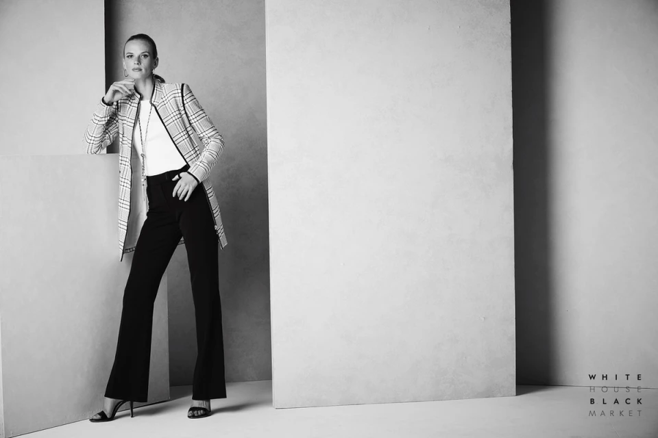 Monochrome fashion shot of an adult woman in a patterned jacket and black slacks leaning against a gray wall in a photo studio.