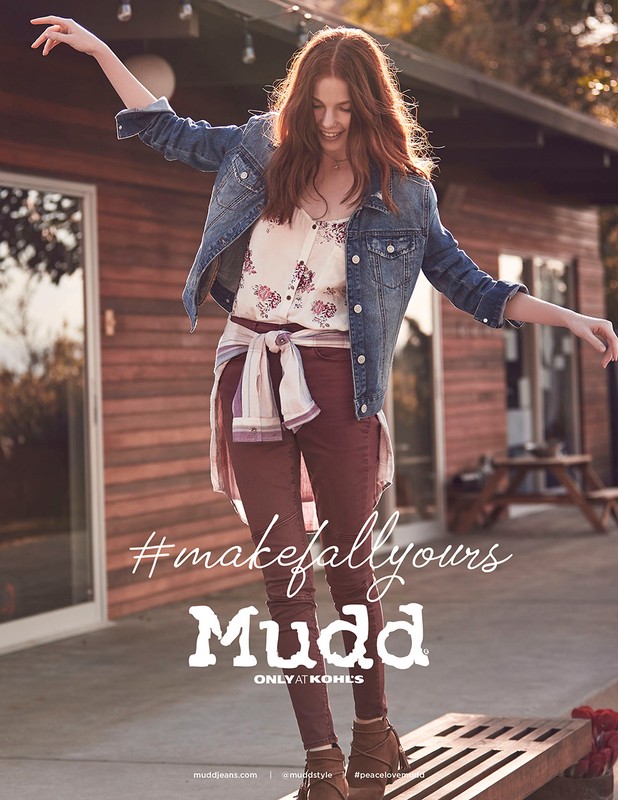 A woman in casual fall fashion poses on a wooden surface outside a wood cabin, showcasing a denim jacket, floral top, and maroon pants.