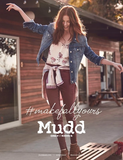 A woman in casual fall fashion poses on a wooden surface outside a wood cabin, showcasing a denim jacket, floral top, and maroon pants.