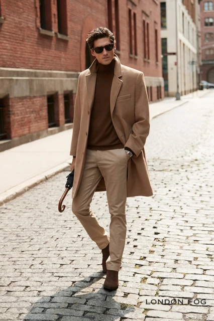 A man in a tan overcoat walks on a cobblestone street, with a building and sky in the background. He's wearing sunglasses and brown boots.