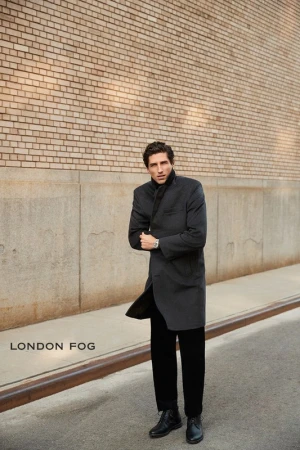 Man wearing a charcoal overcoat standing on a city sidewalk in front of a brick building wall