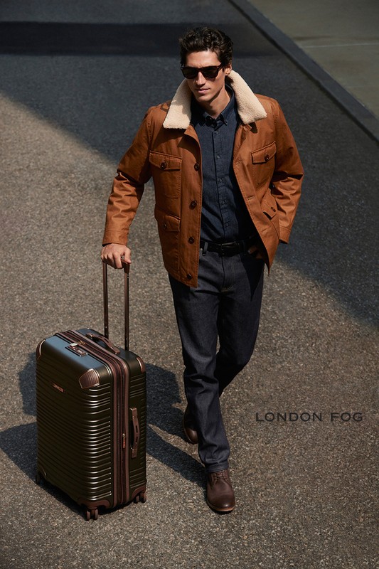 Stylish man in sunglasses walks on city street, pulling an olive suitcase. He wears a brown jacket with fur collar, jeans, and boots.