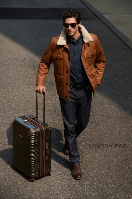 Stylish man in sunglasses walks on city street, pulling an olive suitcase. He wears a brown jacket with fur collar, jeans, and boots.