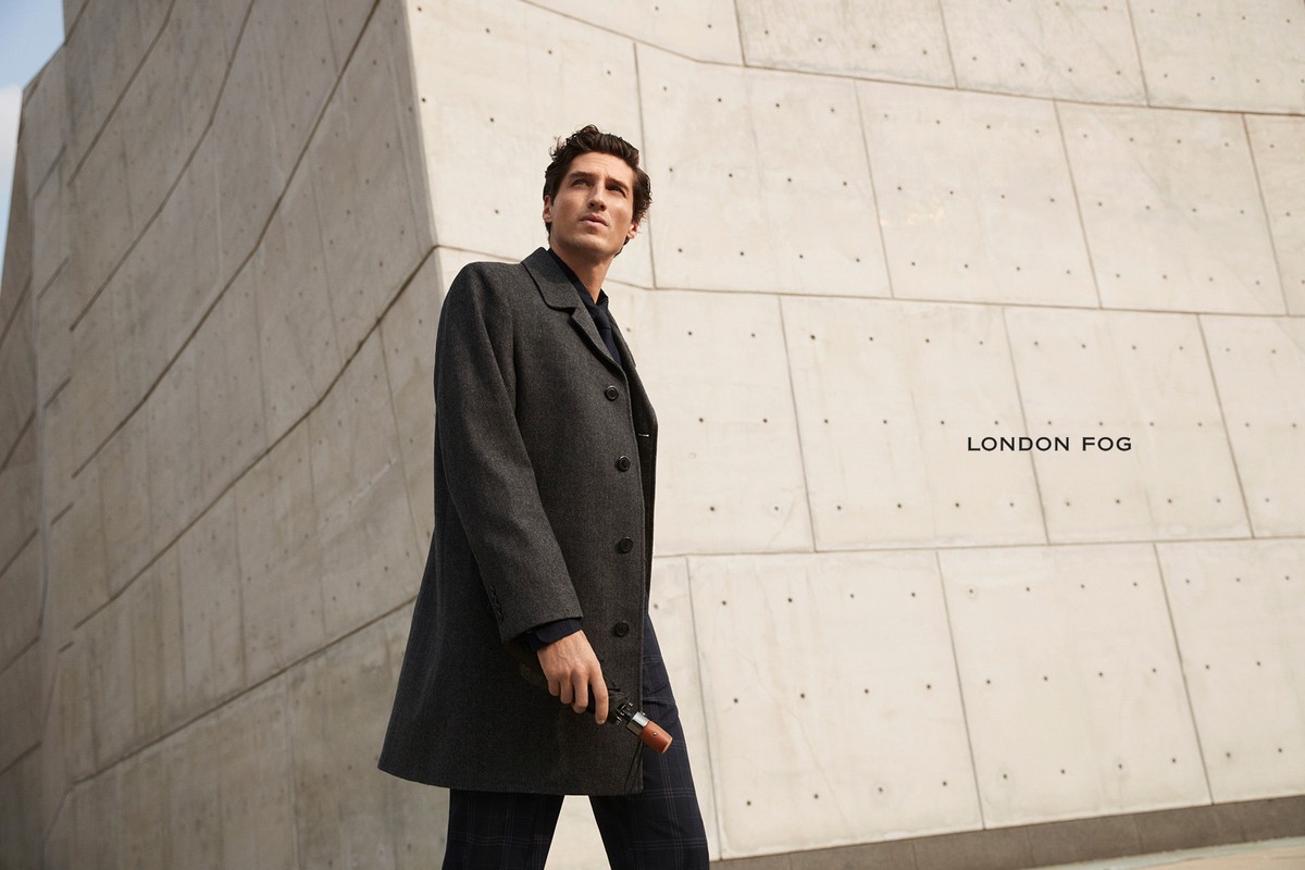 Man wearing a dark grey overcoat holding a folded umbrella against a concrete architectural wall with the text London Fog visible