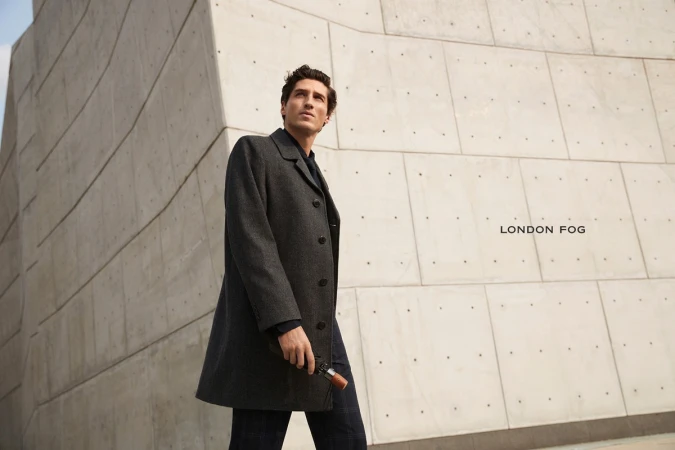 Man wearing a dark grey overcoat holding a folded umbrella against a concrete architectural wall with the text London Fog visible
