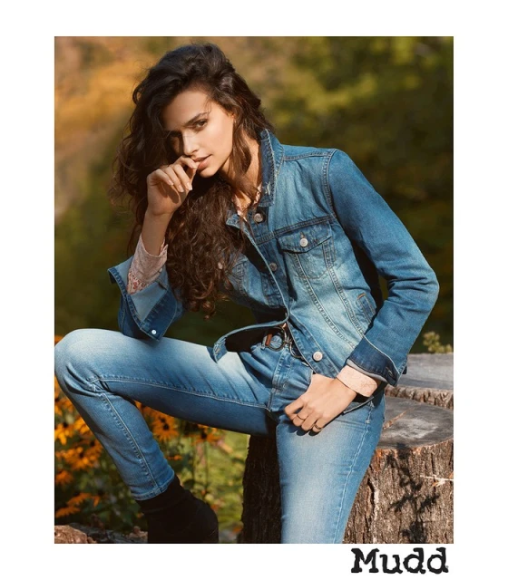 A young woman with long, dark curly hair is wearing a denim jacket and jeans. She's outdoors, seated, and looking at the camera.