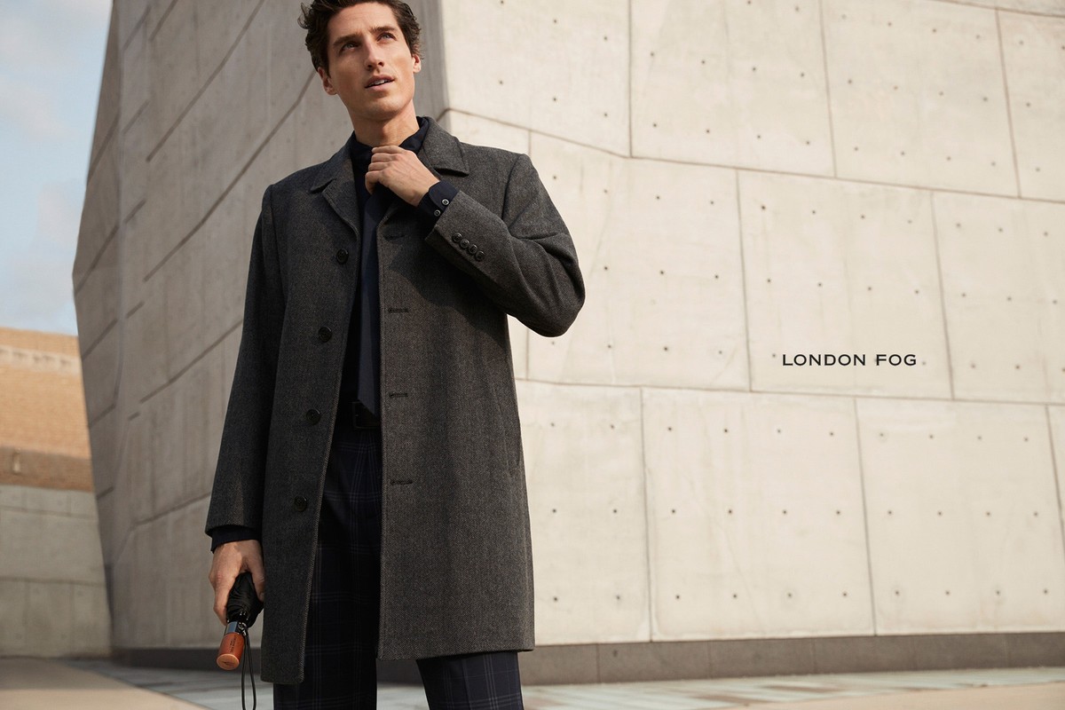 A man in a grey overcoat stands in front of a modern concrete building, holding an umbrella. The image features a clean, commercial aesthetic.