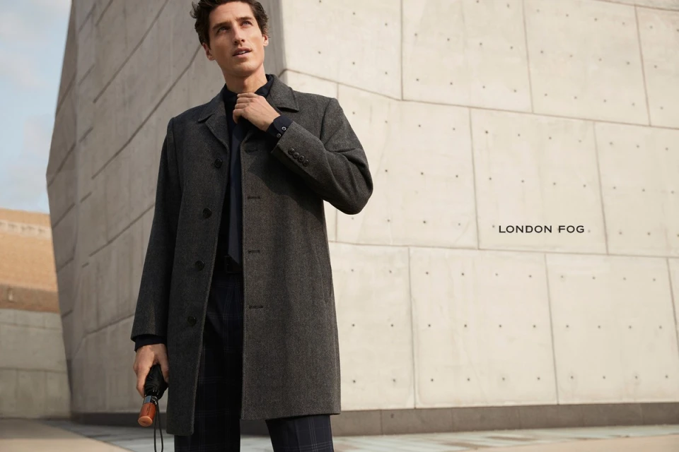A man in a grey overcoat stands in front of a modern concrete building, holding an umbrella. The image features a clean, commercial aesthetic.