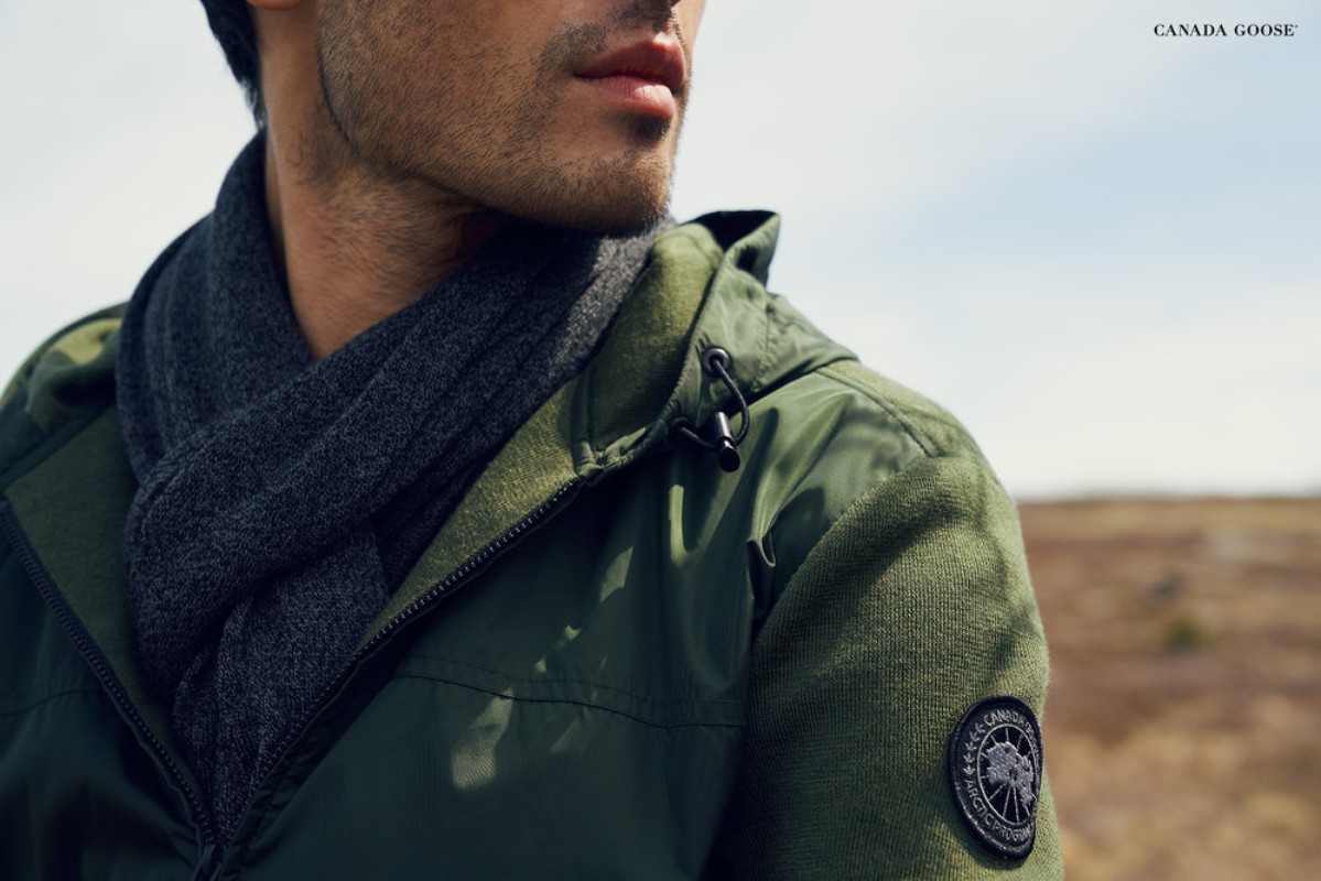 Close-up of a man wearing a green parka and a grey scarf, Canada Goose logo visible on the jacket sleeve. Outdoor setting.