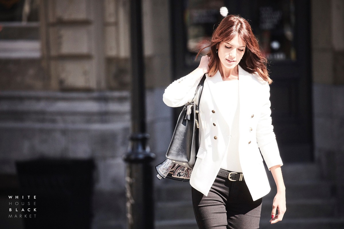 Woman with long brown hair wearing a white double-breasted blazer and black trousers carrying a black handbag