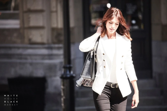 Woman with long brown hair wearing a white double-breasted blazer and black trousers carrying a black handbag