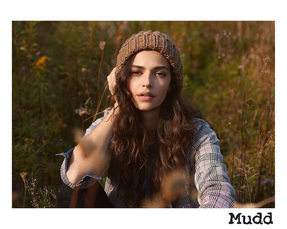 Portrait of a woman with long brown hair wearing a knitted cap and a plaid shirt in a grassy outdoor setting. Soft natural light.
