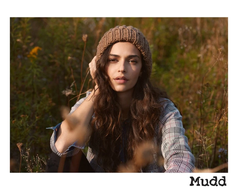 Portrait of a woman with long brown hair wearing a knitted cap and a plaid shirt in a grassy outdoor setting. Soft natural light.