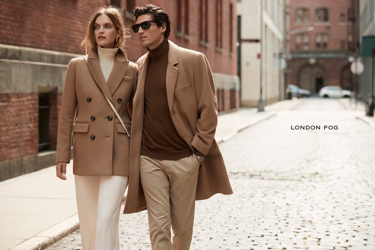 A stylish couple walks on a cobblestone street. The woman wears a camel coat and white pants, while the man is in a brown turtleneck and coat.