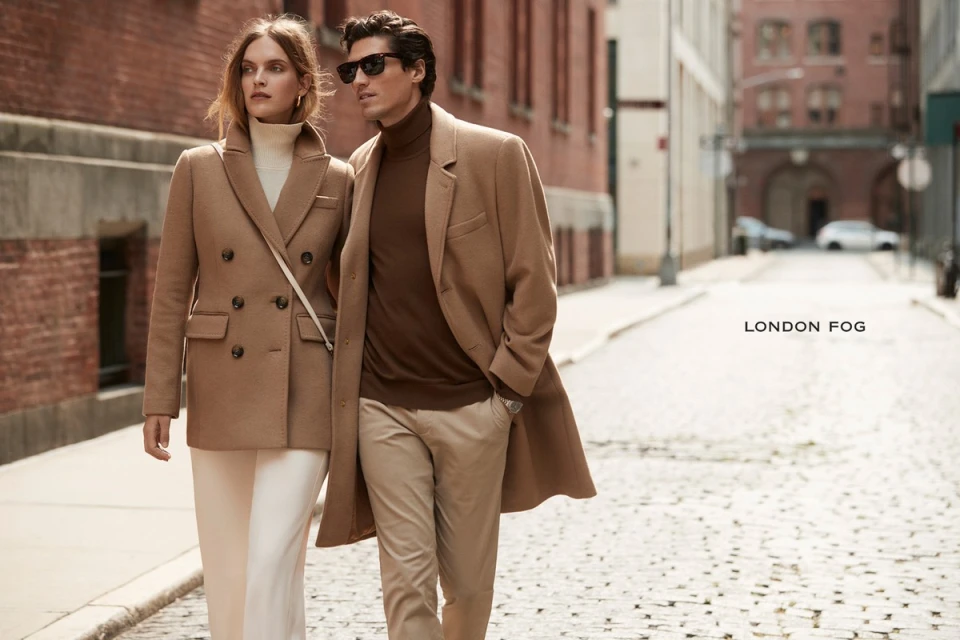 A stylish couple walks on a cobblestone street. The woman wears a camel coat and white pants, while the man is in a brown turtleneck and coat.