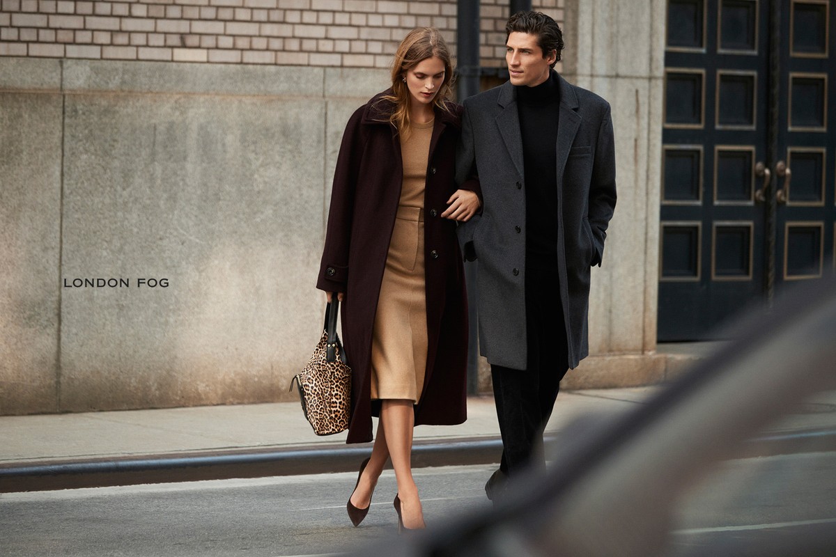 An adult couple walks arm-in-arm on a city sidewalk in a fashion ad; the woman carries an animal-print purse and wears a dark coat and heels.
