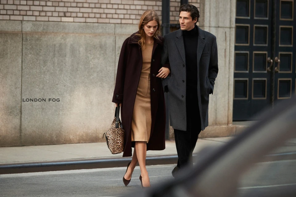 An adult couple walks arm-in-arm on a city sidewalk in a fashion ad; the woman carries an animal-print purse and wears a dark coat and heels.