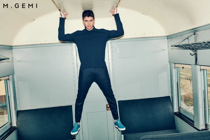Man in dark blue turtleneck and trousers wearing blue sneakers, holding up the ceiling inside a train car