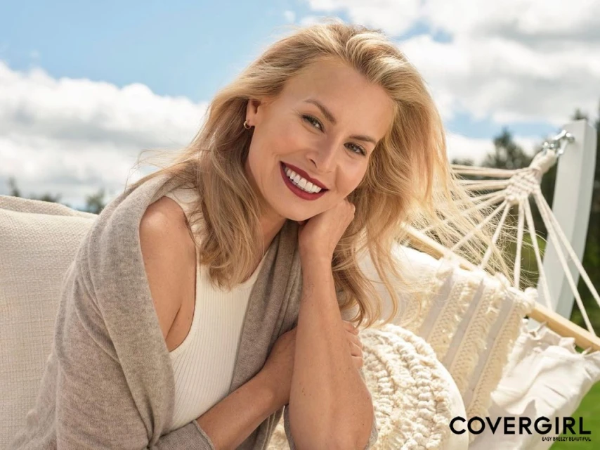 Blonde woman smiles in a hammock, wearing a beige cardigan and white top, with a sunny sky and trees visible in the background.