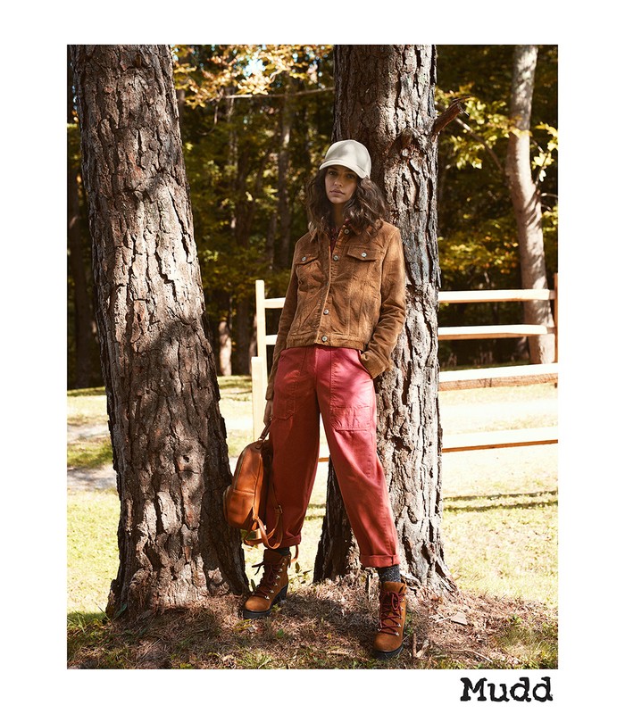 A young woman in outdoor fashion poses by a tree in a forest wearing a corduroy jacket, red pants, and hiking boots with a backpack.