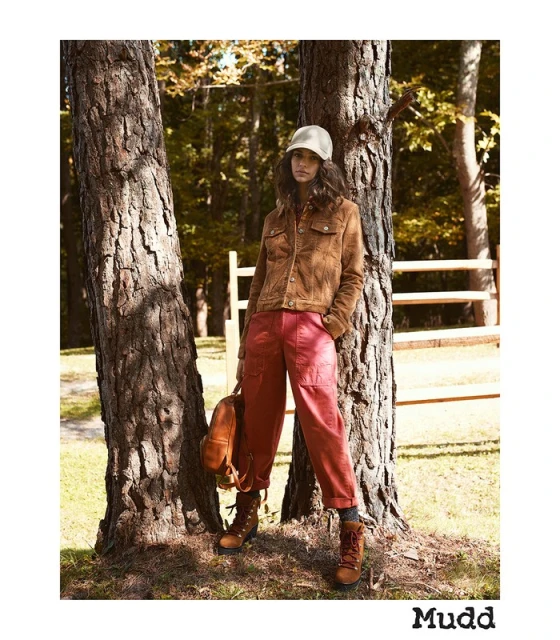 A young woman in outdoor fashion poses by a tree in a forest wearing a corduroy jacket, red pants, and hiking boots with a backpack.