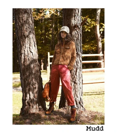 A young woman in outdoor fashion poses by a tree in a forest wearing a corduroy jacket, red pants, and hiking boots with a backpack.