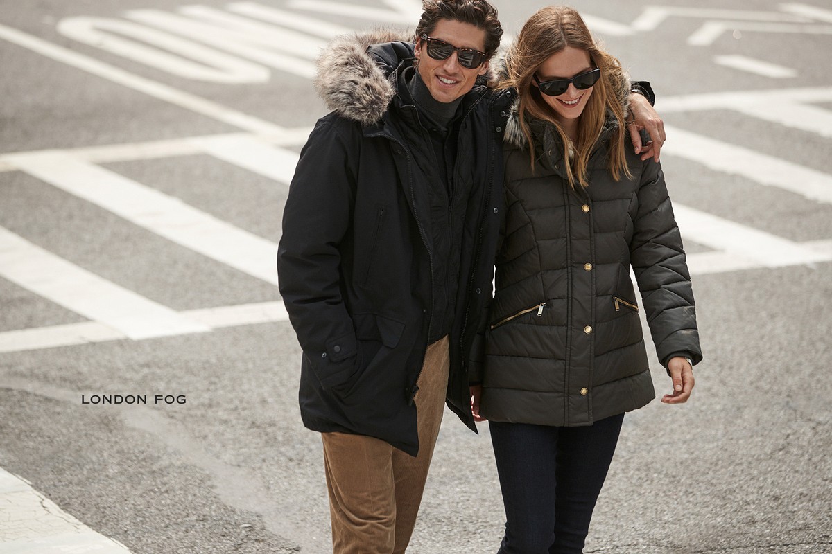 A stylish couple in winter coats walks on an urban street. The man's black parka features a fur-lined hood; the woman's quilted coat is dark green.