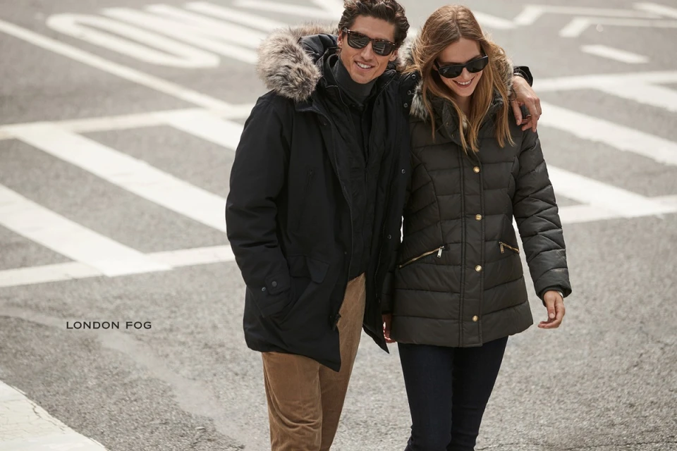 A stylish couple in winter coats walks on an urban street. The man's black parka features a fur-lined hood; the woman's quilted coat is dark green.