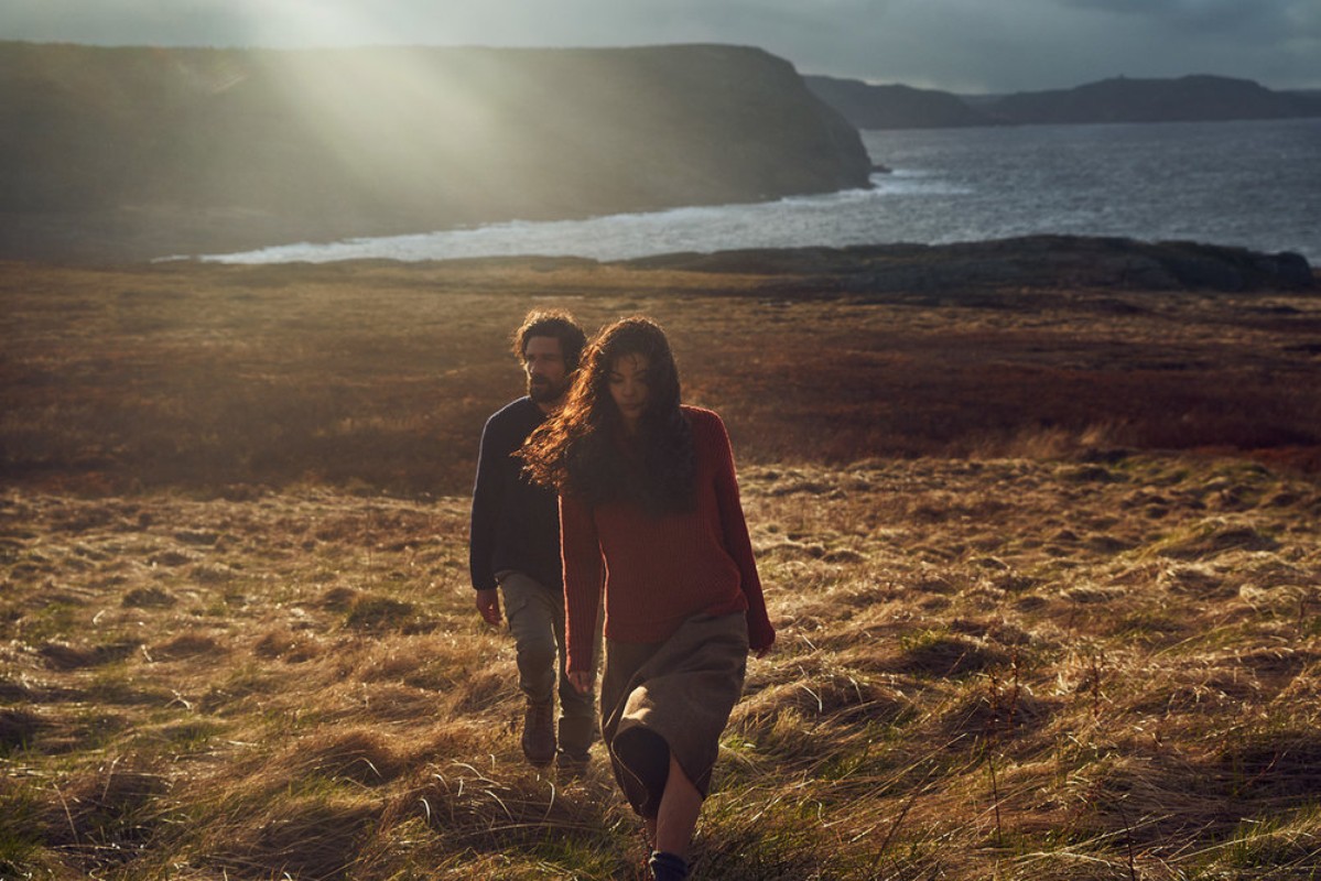 Couple walks through grassy field near the ocean on a cloudy day with golden light. Landscape includes cliff and hazy sunlight.