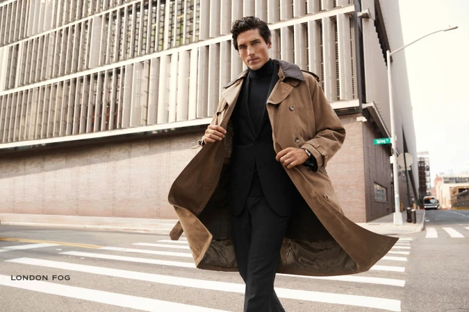 Man wearing a camel trench coat over a black suit and turtleneck walking across a city street.