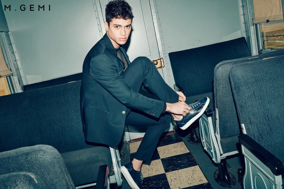 An adult male tying his shoelaces while seated on a train. He wears a suit and sneakers and the scene is dimly lit.