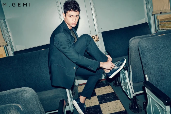 An adult male tying his shoelaces while seated on a train. He wears a suit and sneakers and the scene is dimly lit.
