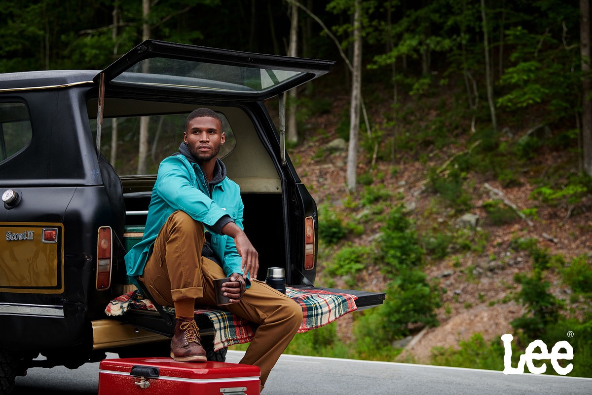 Adult male sits on the back of an SUV in front of a forest wearing a turquoise jacket, brown pants and boots, and holding a mug.