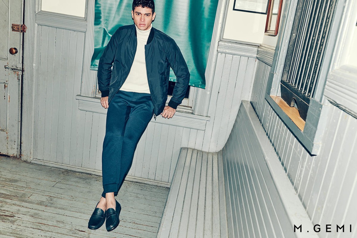 A young adult male in a dark bomber jacket and blue pants sits on a gray wooden bench in an indoor setting, fashion portrait.