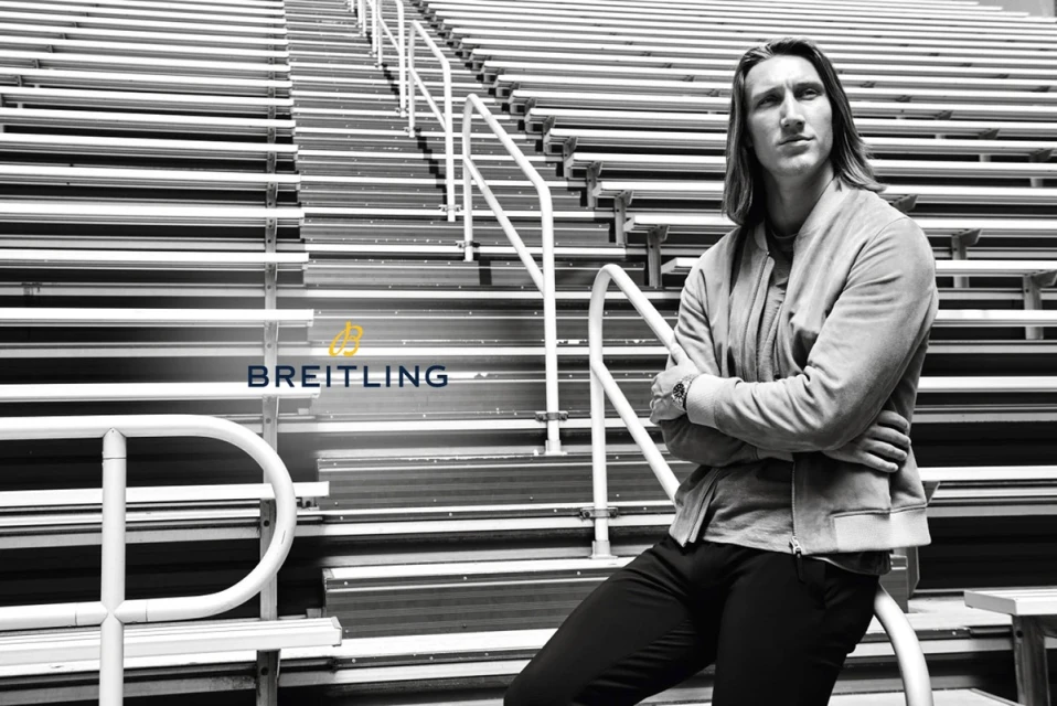 Adult male with long hair wearing a jacket sitting on stadium bleachers in black and white with watch and Breitling logo.