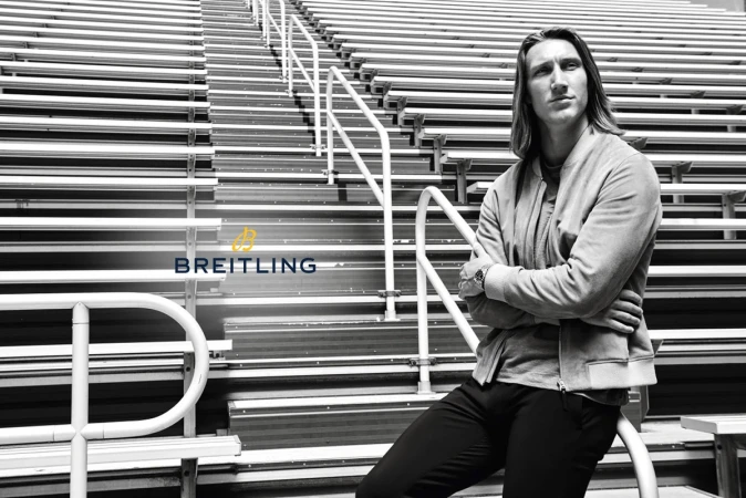Adult male with long hair wearing a jacket sitting on stadium bleachers in black and white with watch and Breitling logo.
