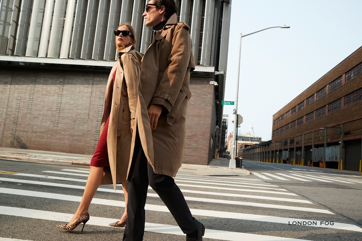 A stylish couple in trench coats crossing a city street, showcasing fashion against an urban backdrop with zebra stripe crosswalk.